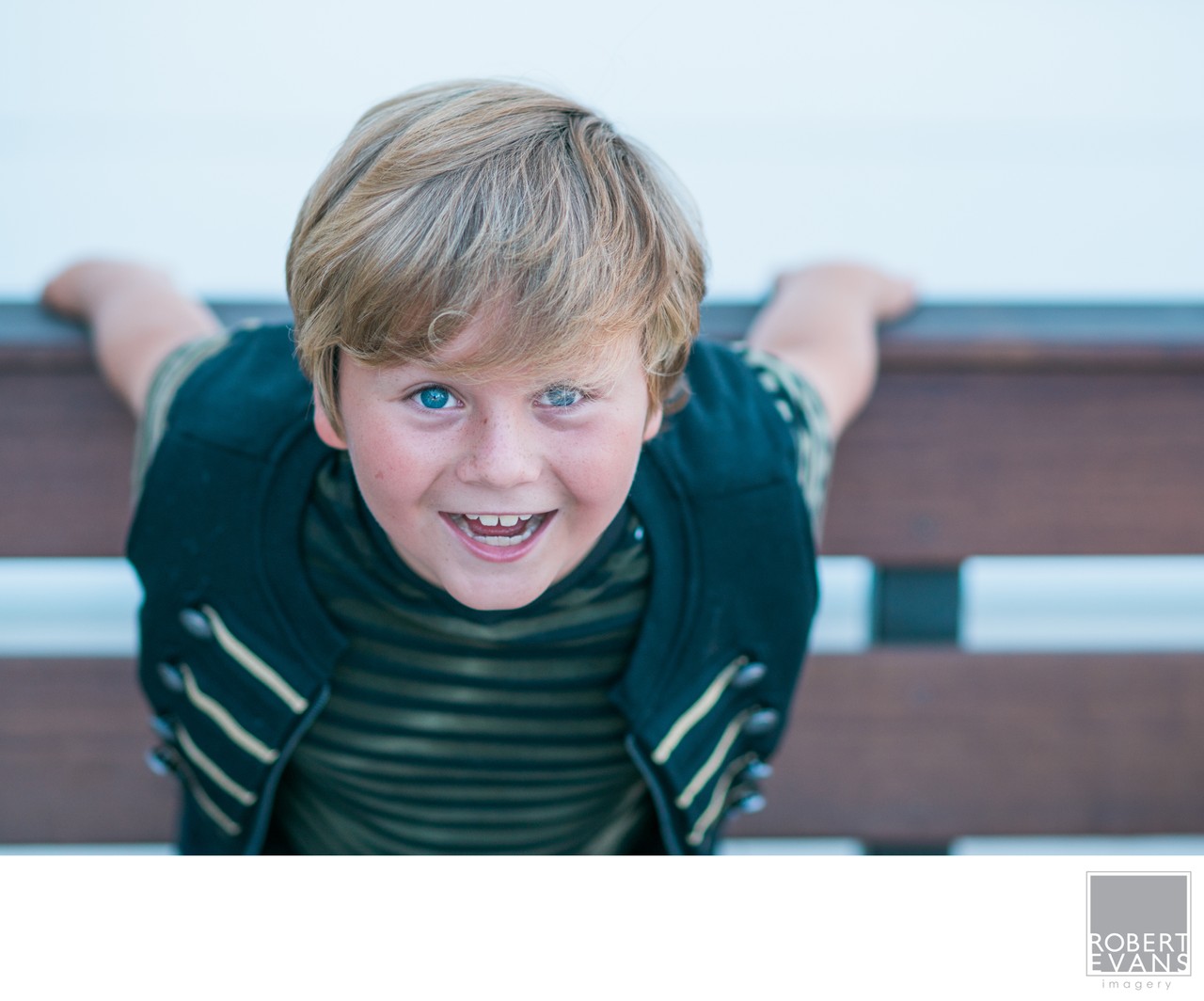 A smiling boy in a striped shirt leans toward the camera over the back of a bench.