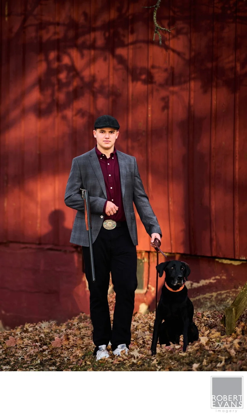 A young man in hunting clothes poses with a dog and rifle beside a red barn.