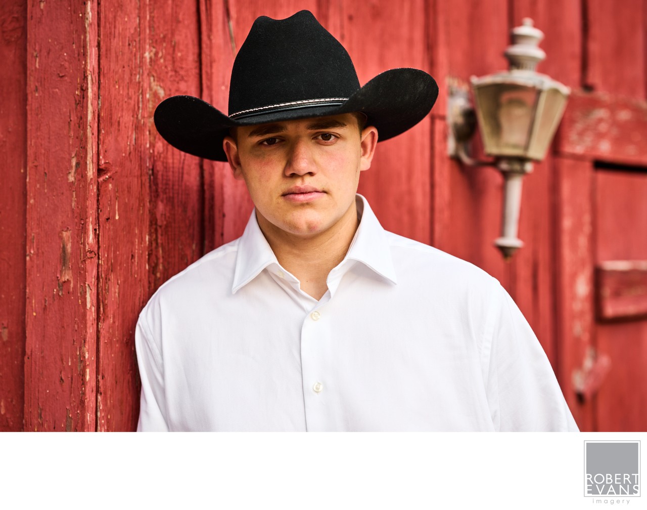 A young man in a black cowboy hat and white shirt stands beside a red barn.