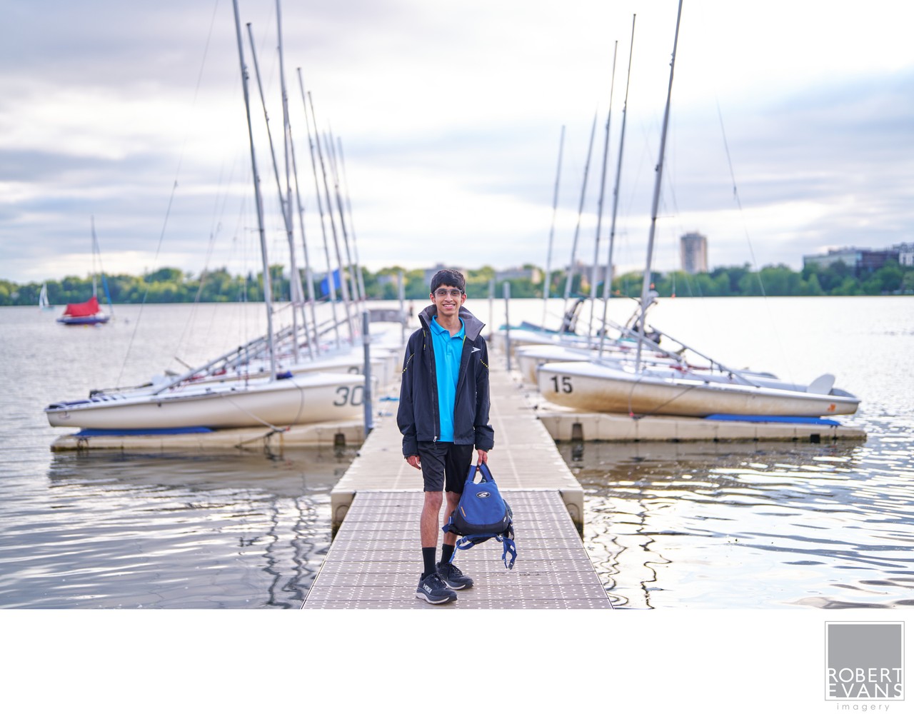 A young man stands on a dock with sailboats behind him.