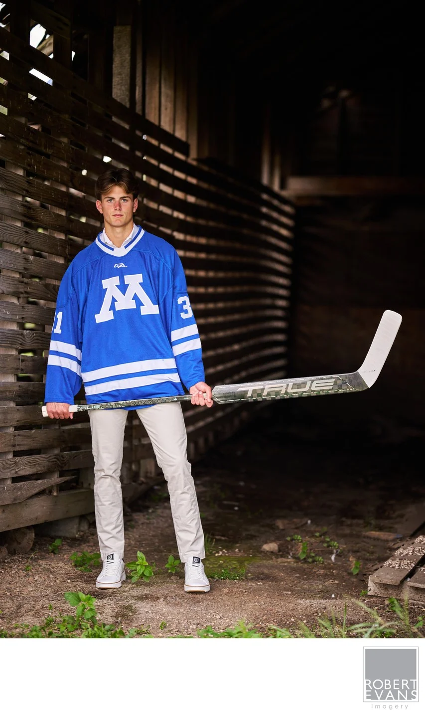 A young man in a blue hockey jersey holds his stick beside a dark barn.