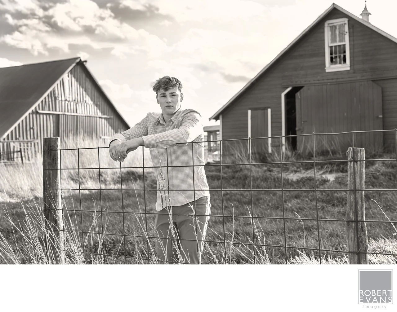 A young man leans against a wire fence between farm buildings in black and white.