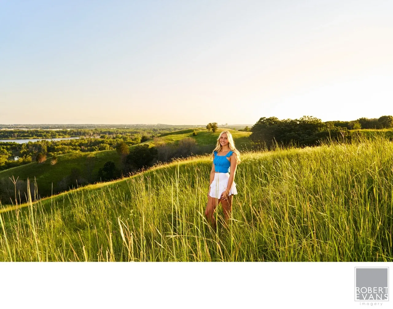 A young woman stands in tall grass beneath a warm evening sky.