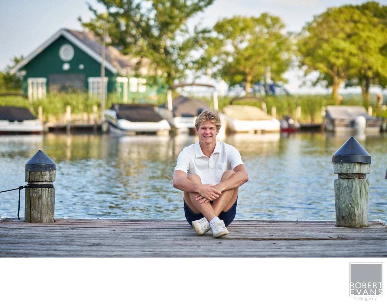 A young man sits cross-legged on a dock beside the water.