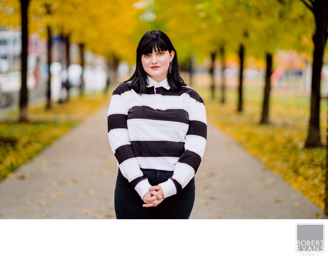 A young woman in a striped sweater stands beneath colorful autumn trees.