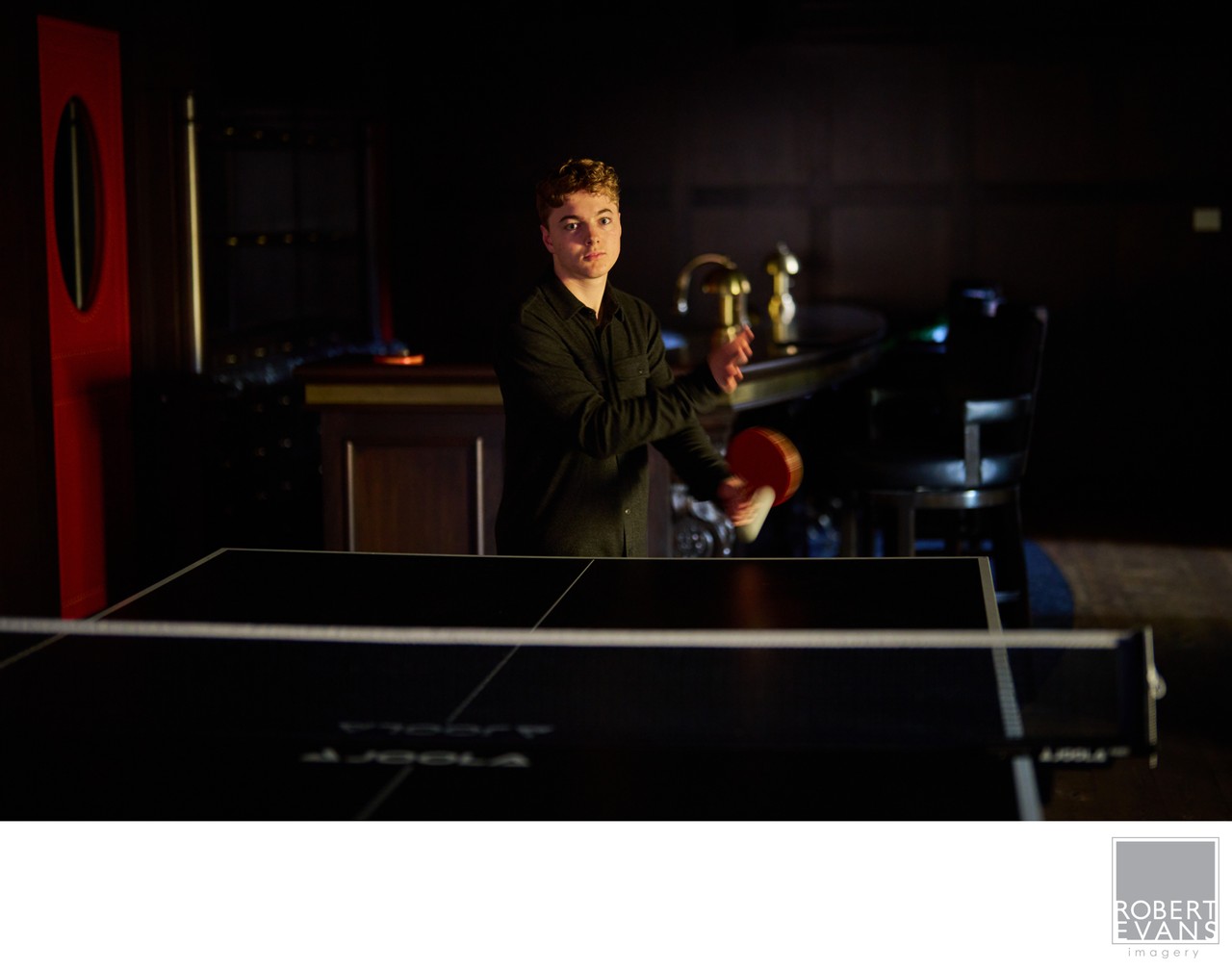 A young man holds a paddle beside a table tennis table at home.