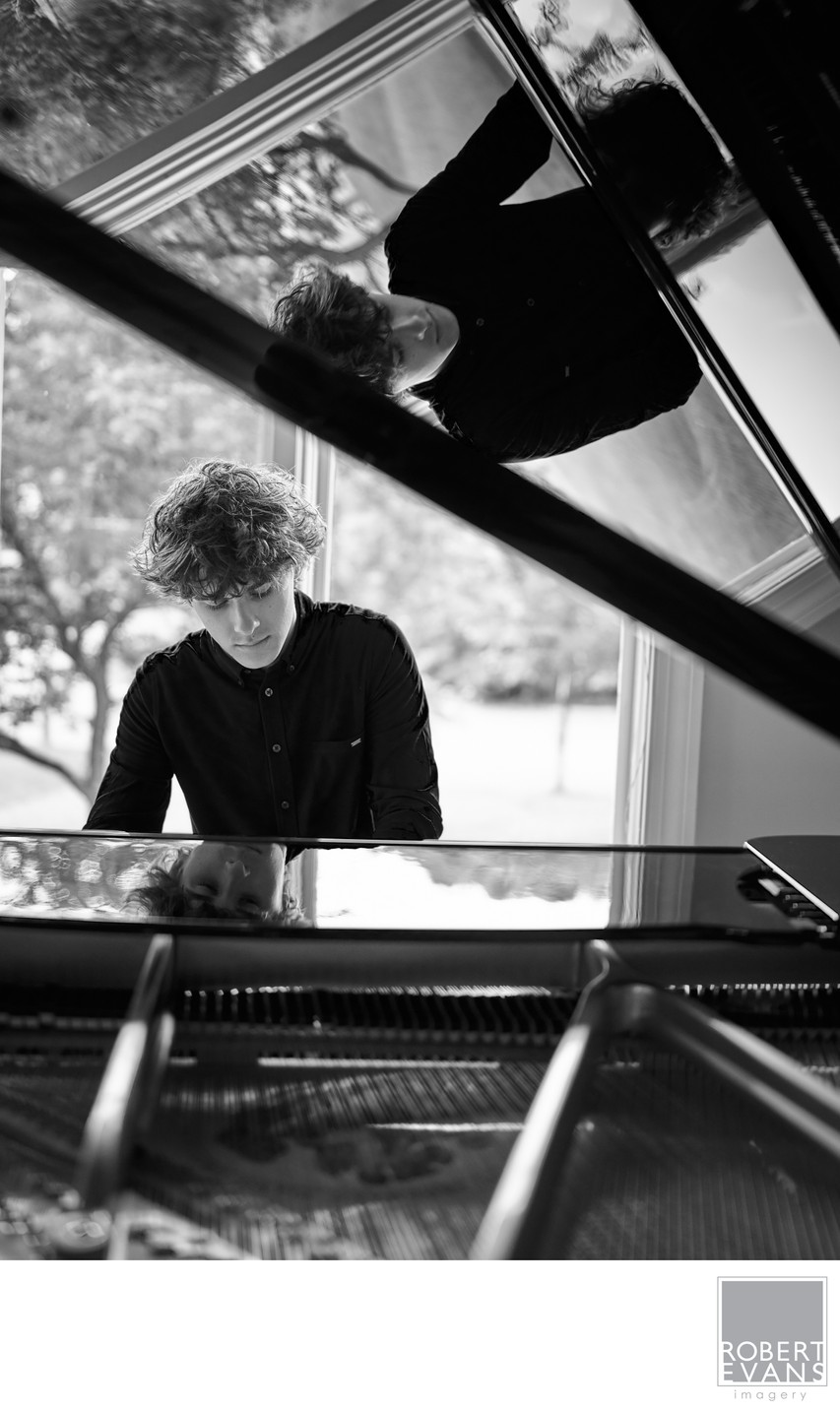 A young man's face is reflected in the open piano lid in black and white.
