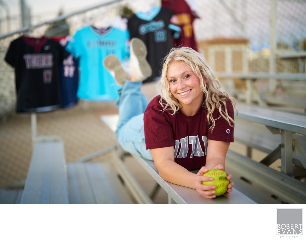A young woman holds a softball while seated in bleachers with jerseys behind her.