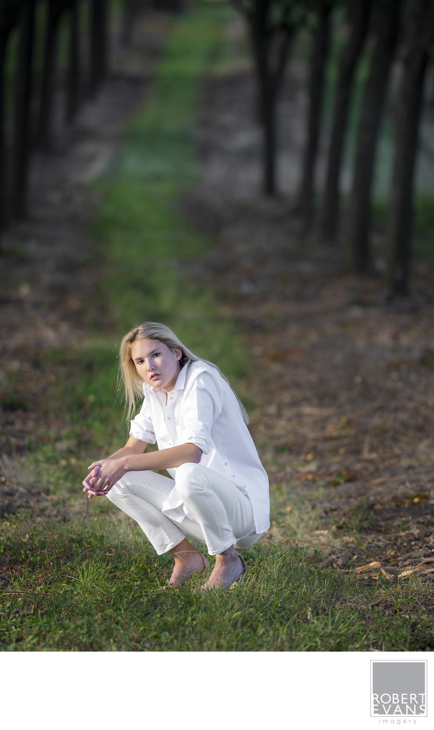 A young woman in a white dress crouches among woodland plants.