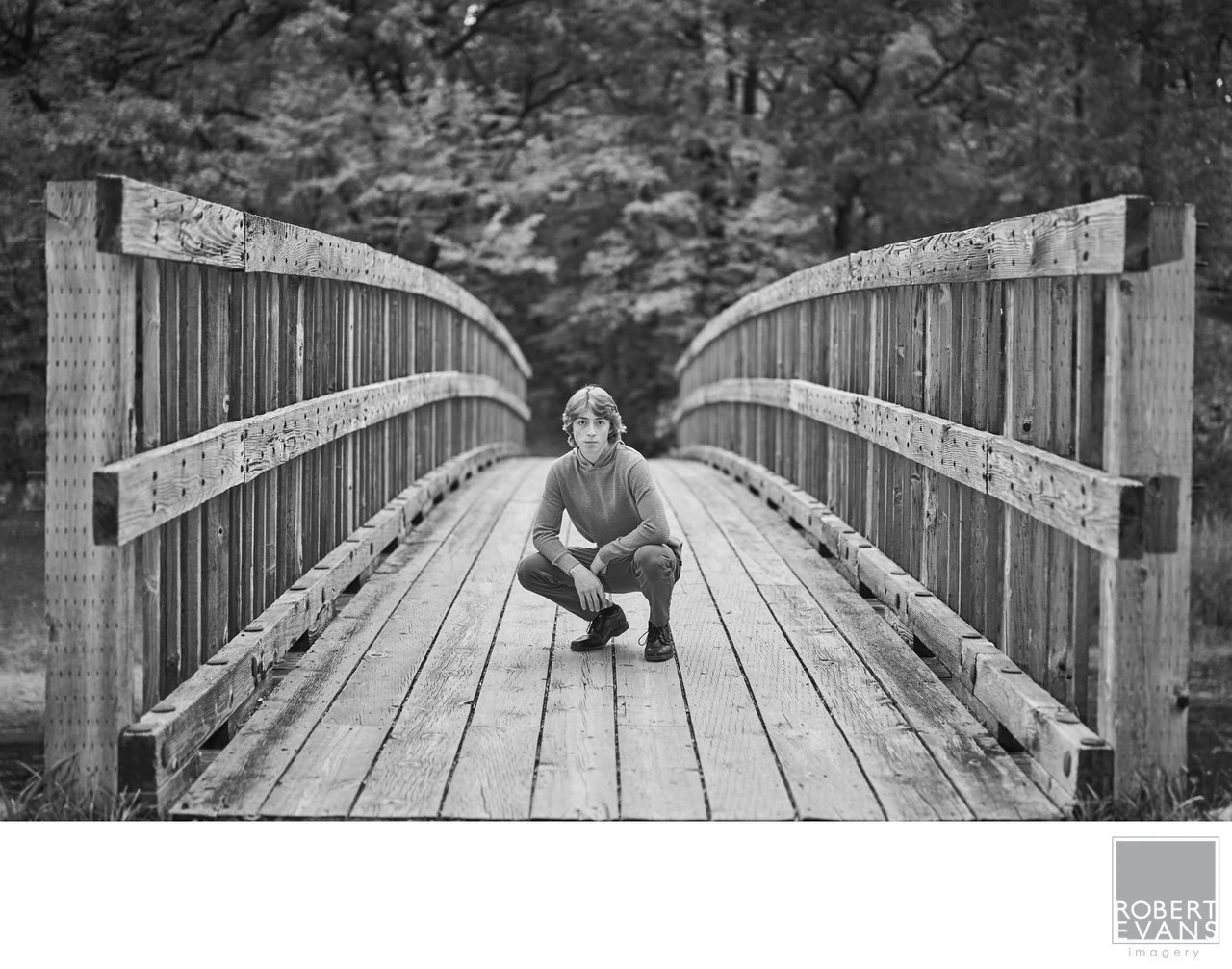 A young man crouches on a wooden footbridge in black and white.
