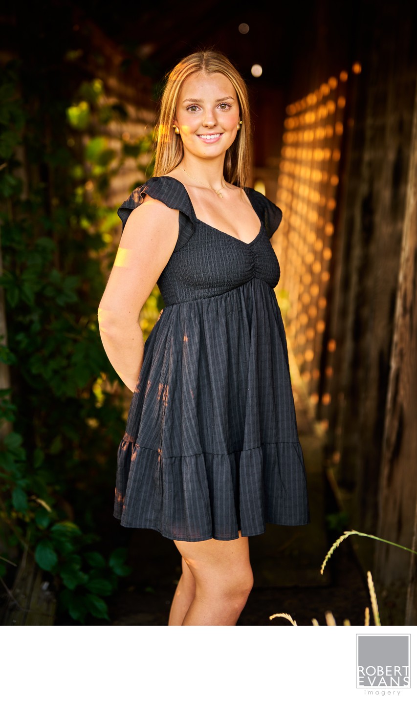 A young woman in a black dress stands between greenhouse doors.