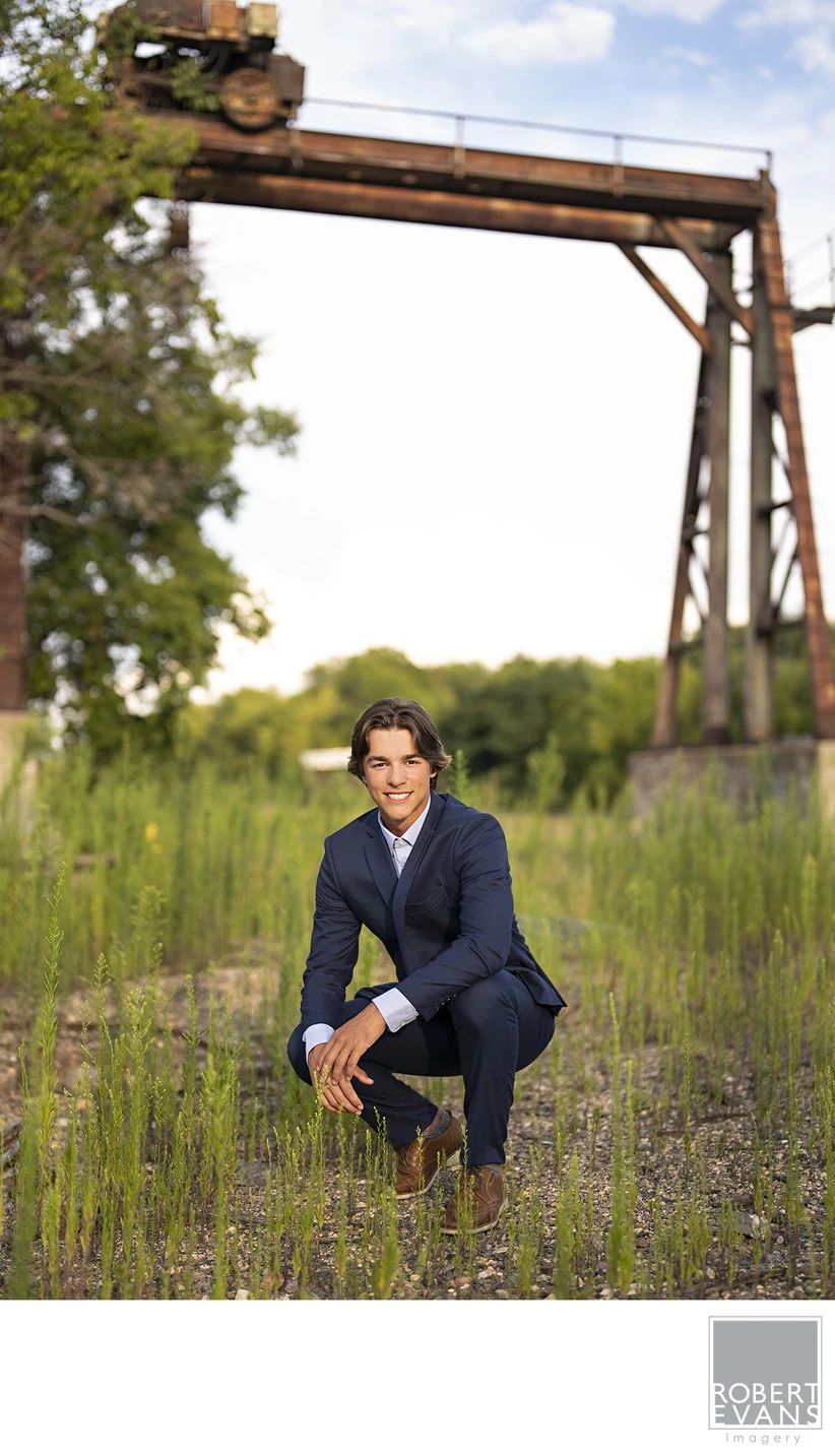 A young man in a suit crouches beside a wooden garden structure.