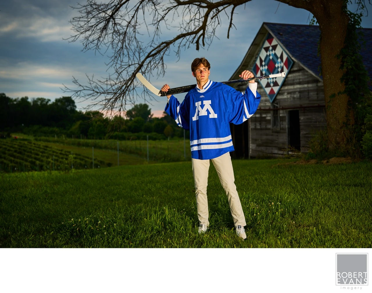 A young man in a blue hockey jersey rests a stick across his shoulders beside a barn.