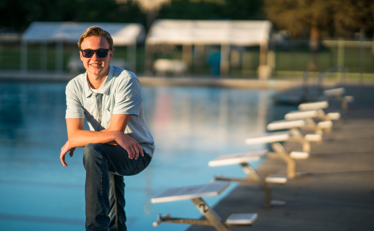A young man wearing sunglasses poses at swimming pool starting blocks.