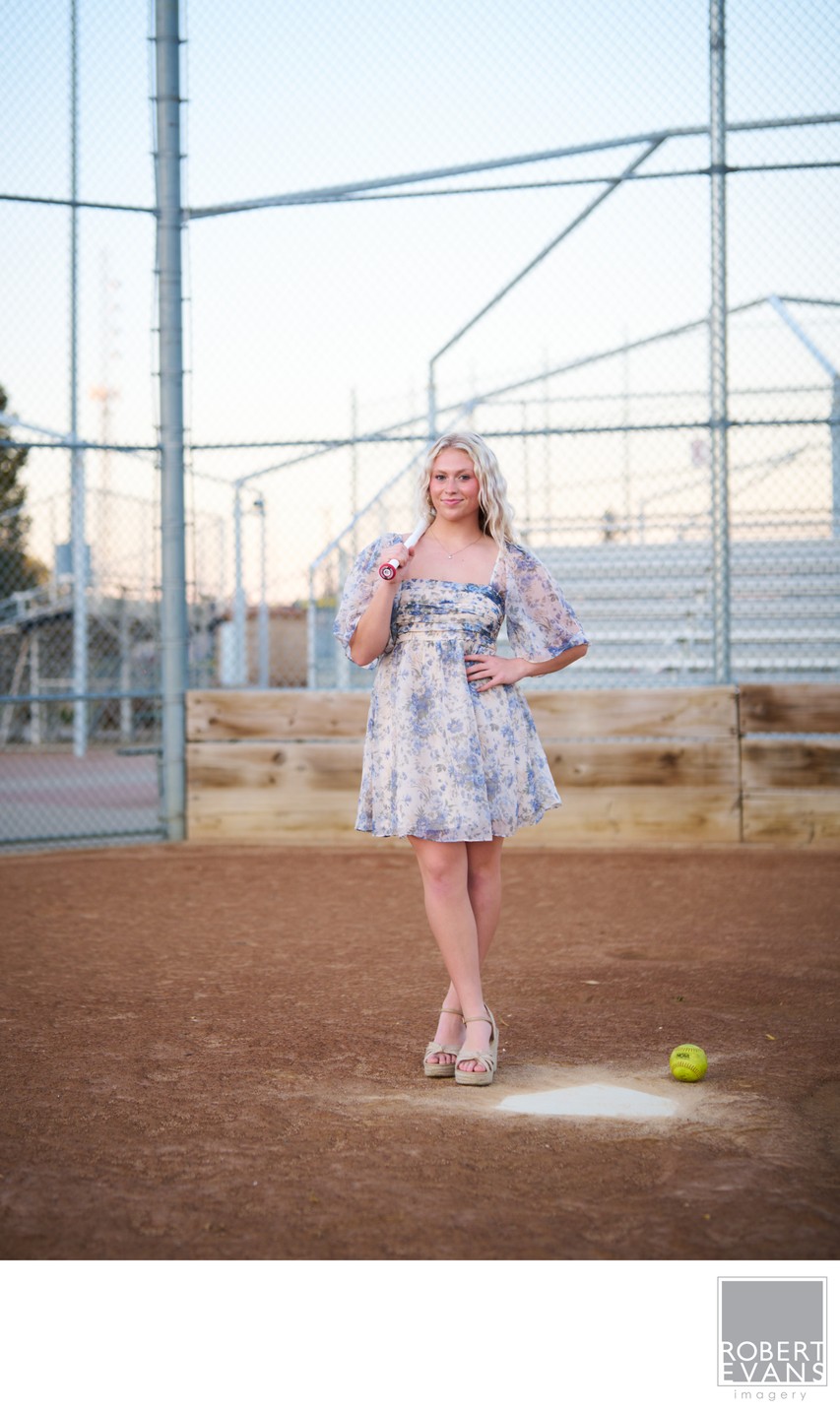 A young woman in a floral dress stands on a softball field with a ball at her feet.