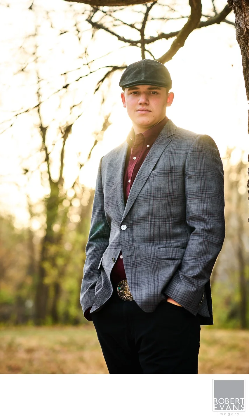 A young man in a blazer and newsboy cap stands among trees.