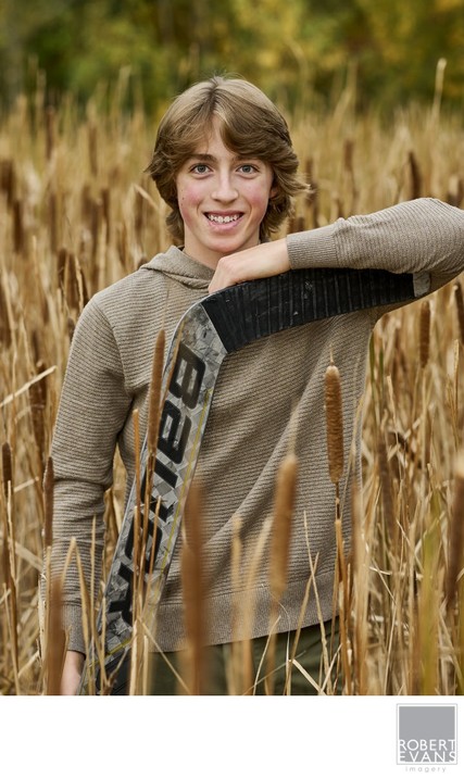 A young man holds a goalie stick beside tall reeds.