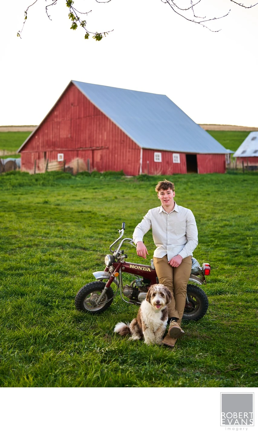 A young man poses with a motorbike and dog near a red barn.
