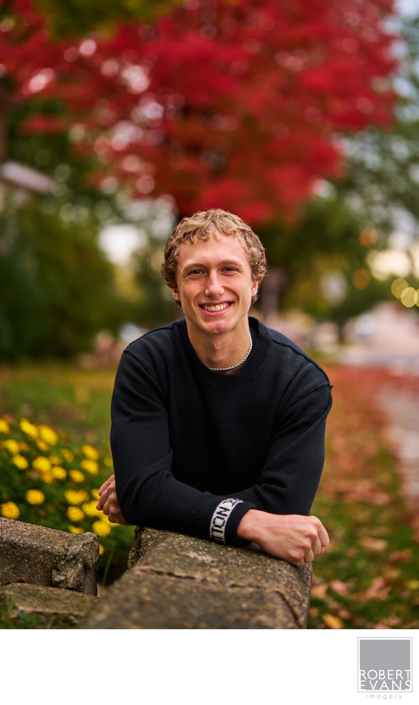 A young man in a black sweater poses against green and red autumn foliage.