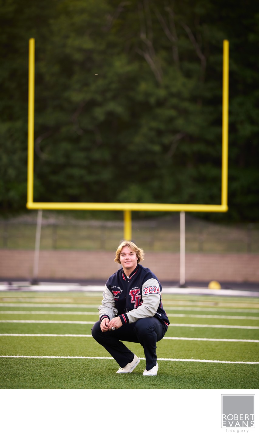 A young man in a varsity jacket crouches beneath football goalposts.