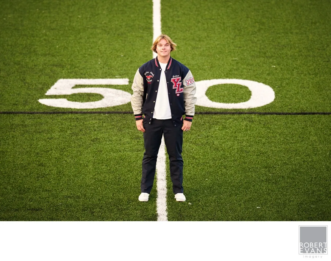 A young man in a varsity jacket stands at the fifty-yard line on a football field.