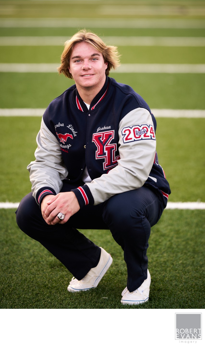 A young man wearing a varsity jacket crouches on the grass.
