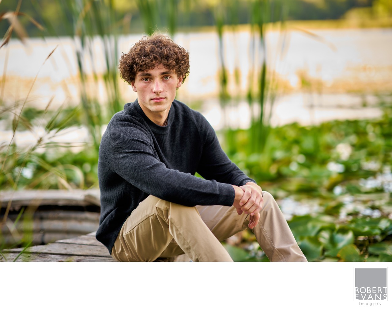 A curly-haired young man sits on a dock beside water and reeds.