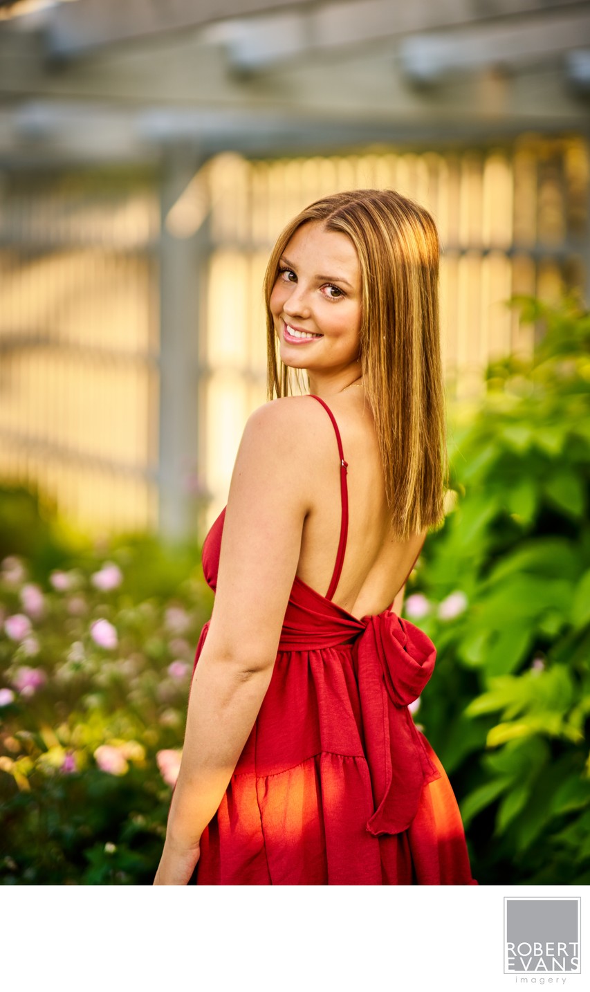 A young woman in a red dress looks back beside a glass greenhouse.