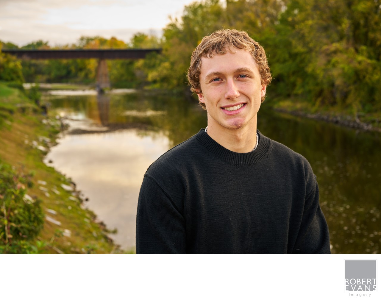 A smiling young man in a black shirt poses beside a creek.