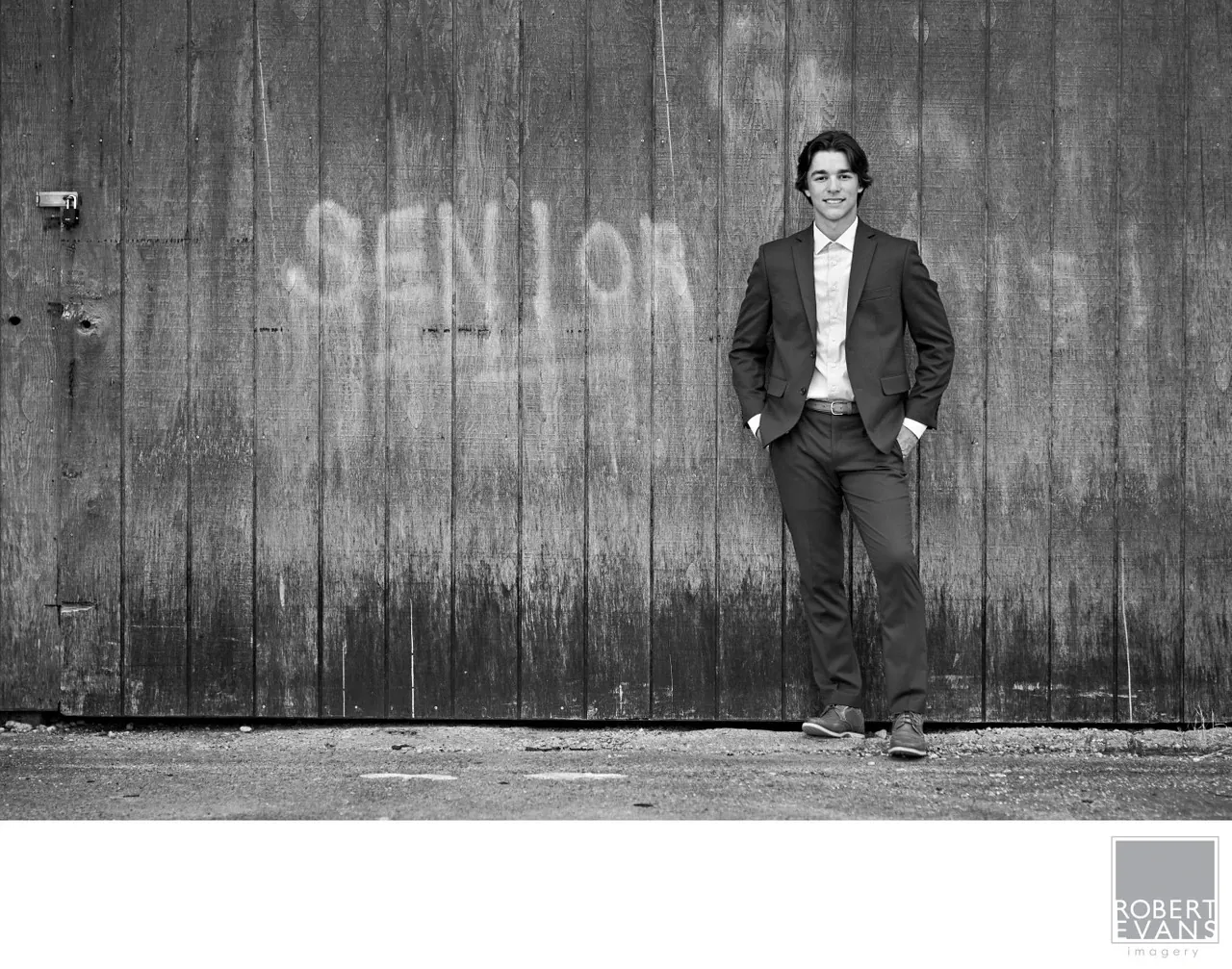 A young man in a suit poses beside a dark wall painted with the word senior, in black and white.