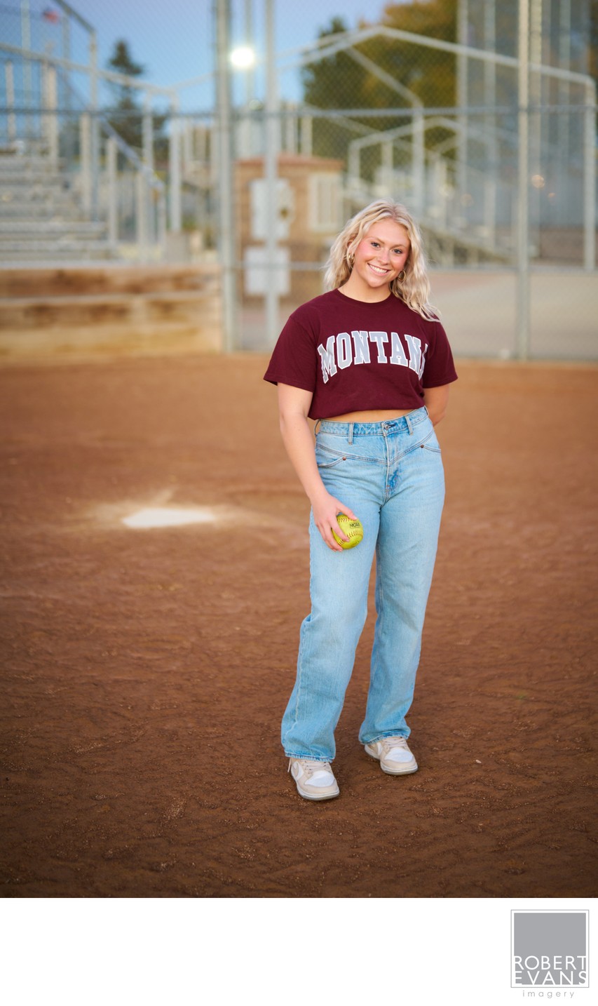 A young woman wearing a Montana shirt holds a softball on a ball field.