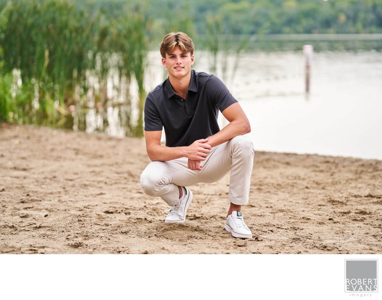 A young man in a black polo shirt crouches beside the beach.