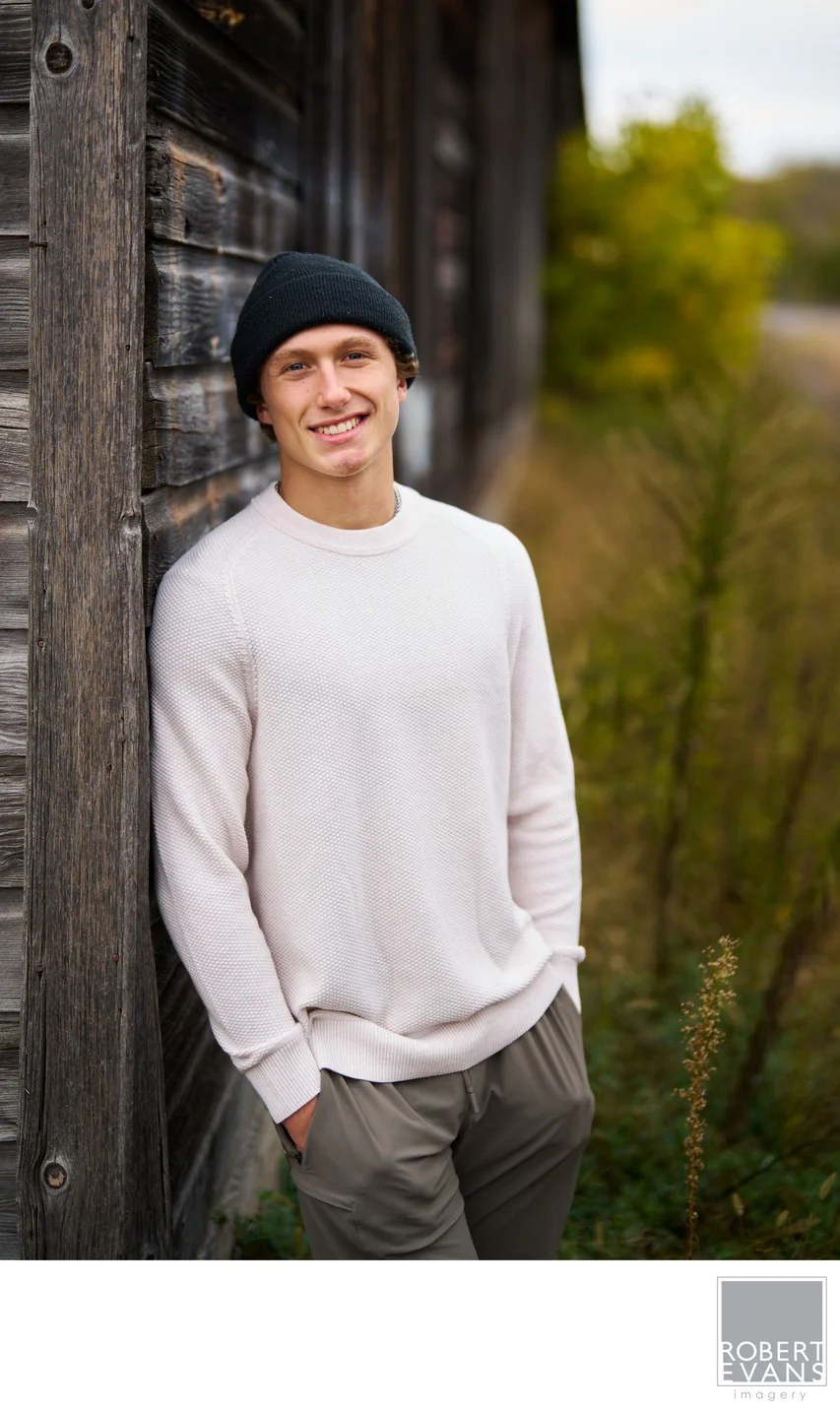 A young man in a cream shirt and dark knit cap poses beside a weathered wall.