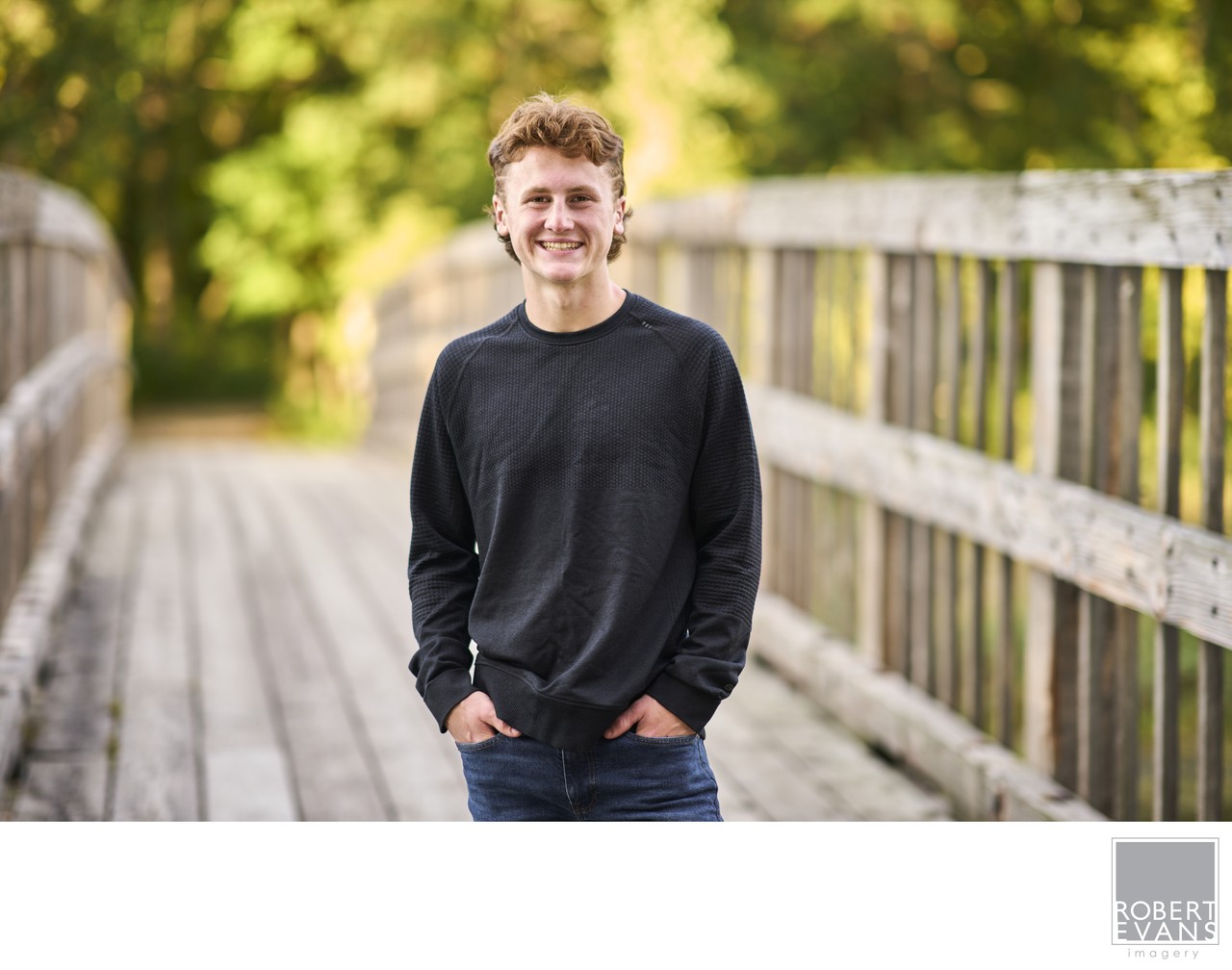A young man in a black sweater stands on a wooden footbridge.