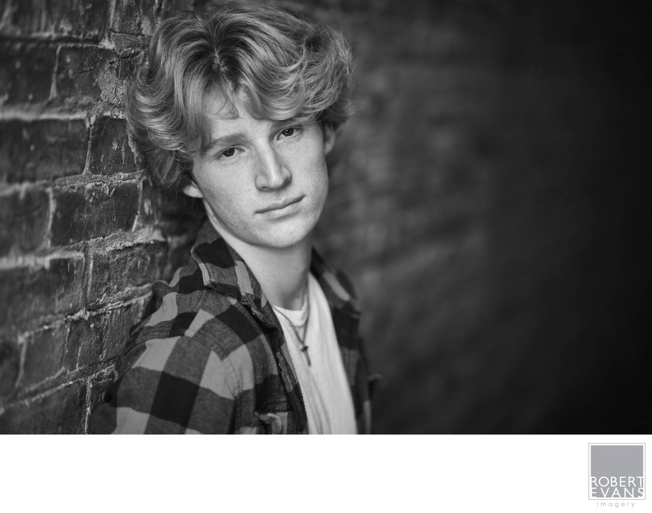 A young man wearing plaid stands beside a brick wall in black and white.