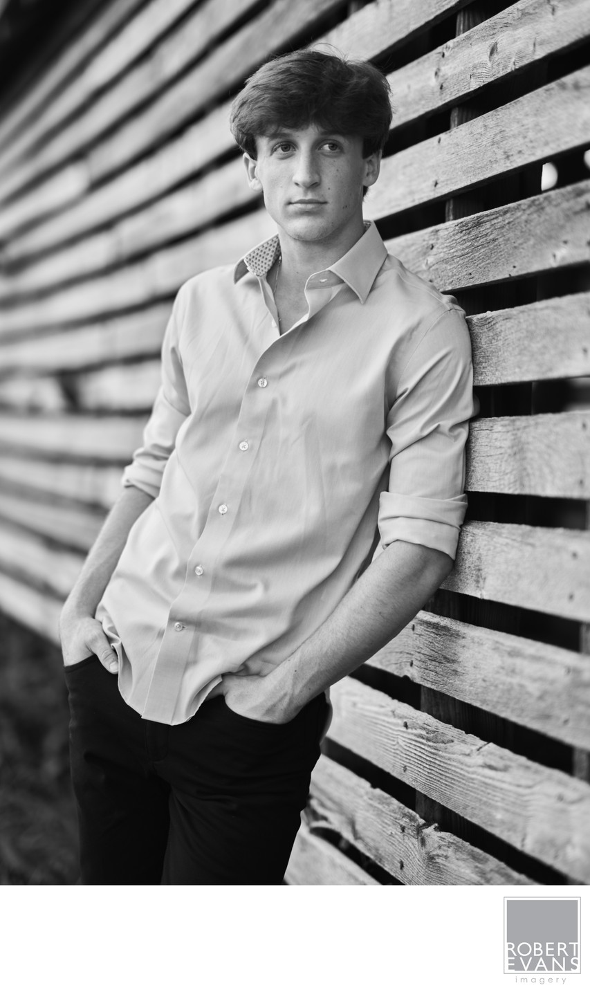 A young man in a light shirt poses beside wooden slats in black and white.