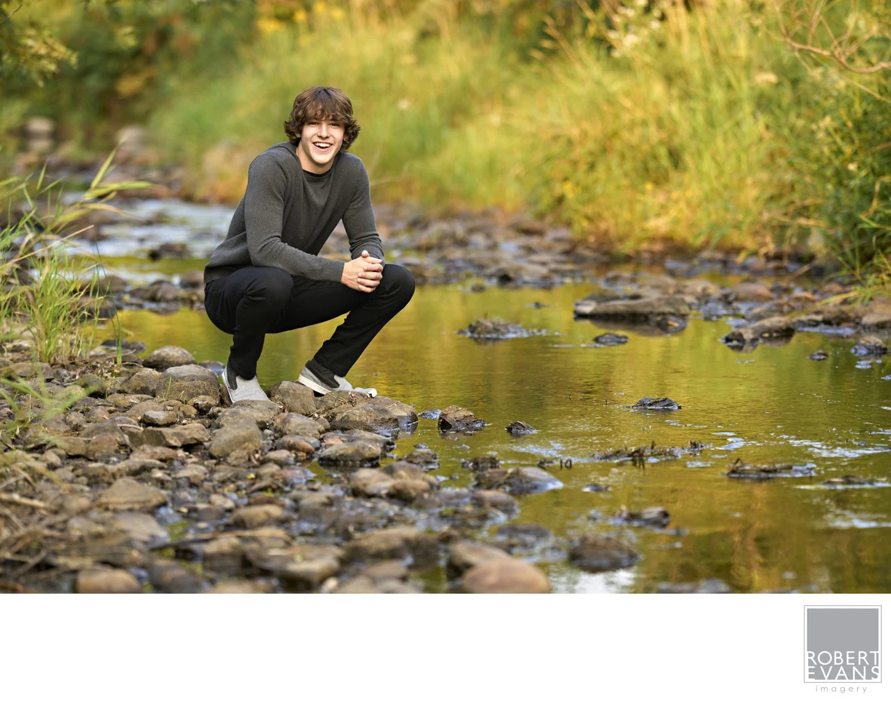 A young man dressed in black crouches on rocks beside a creek.