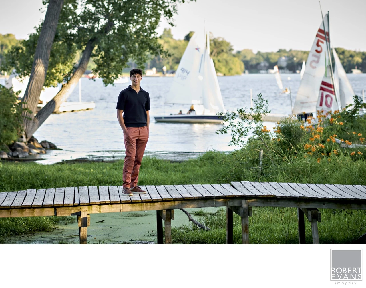 A young man stands on a dock with sailboats in the background.