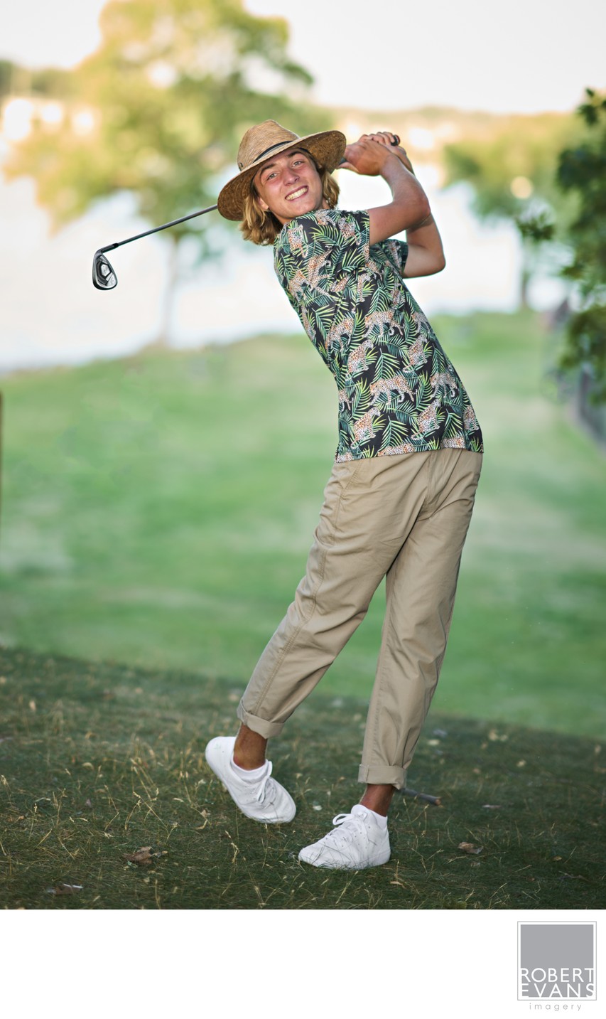 A young man swings a golf club on a course bordered by trees.