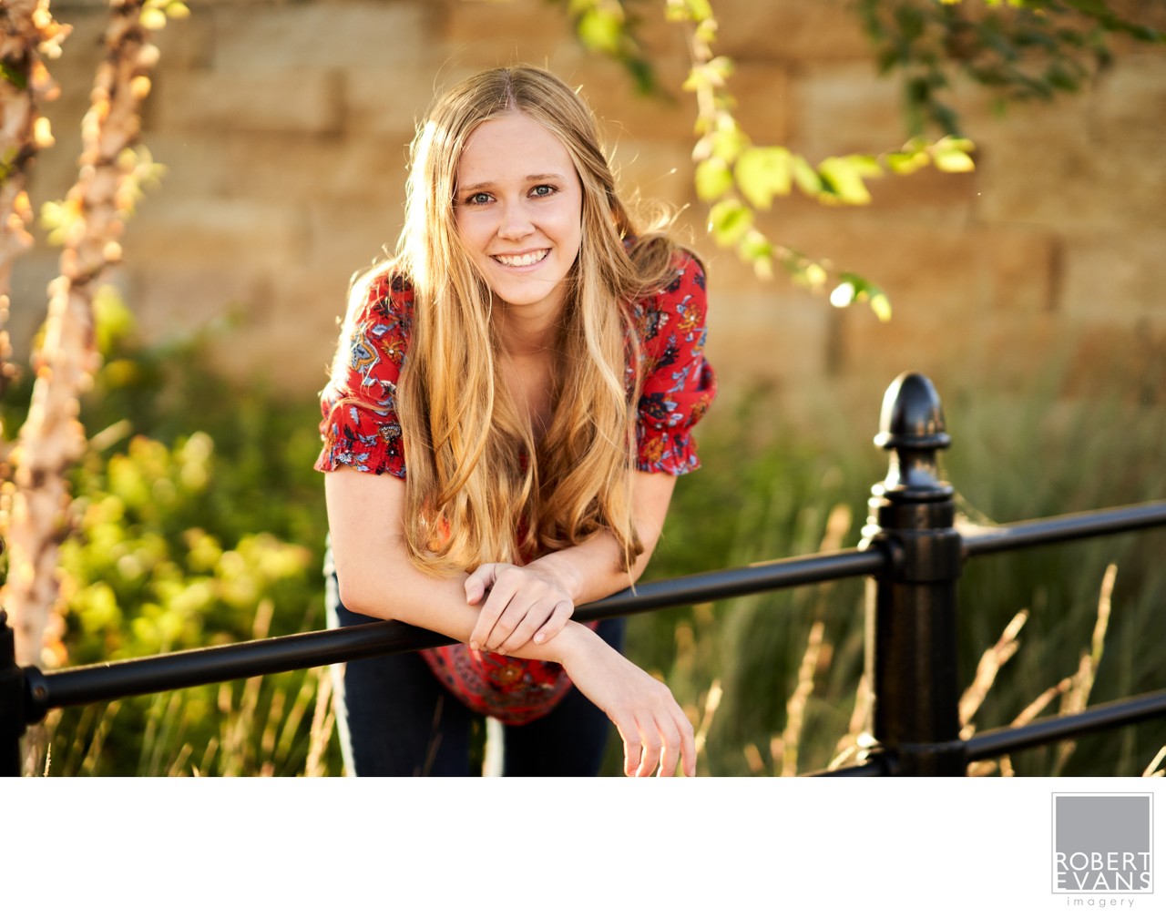 A young woman in a red floral top leans against a black iron fence.