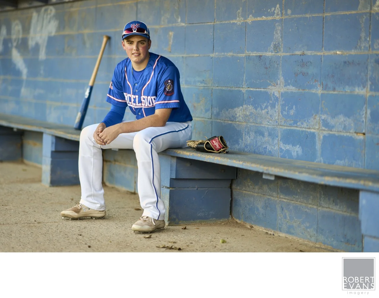 A young baseball player holds a bat in a blue dugout.