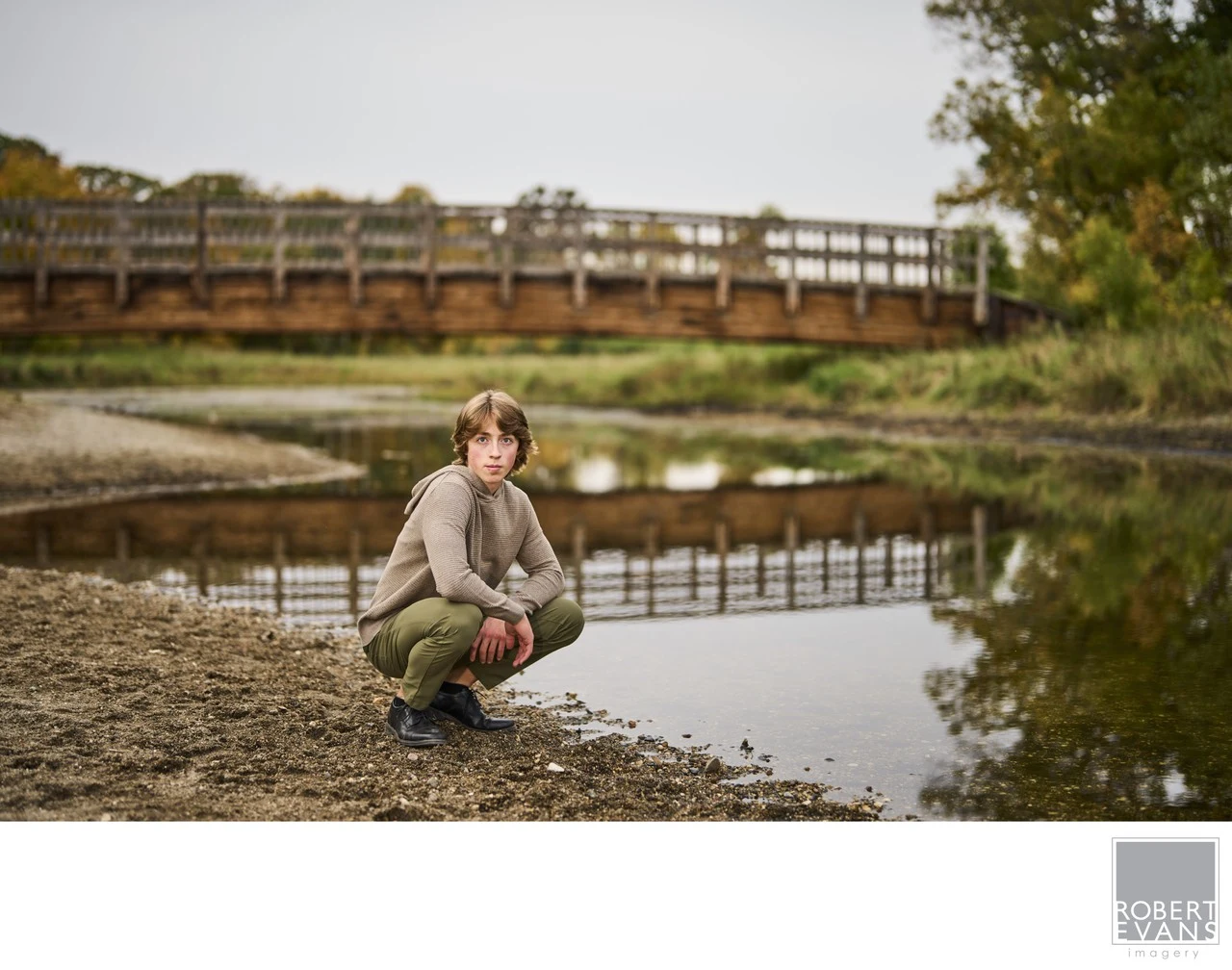 A young man crouches beside a pond reflecting a bridge.