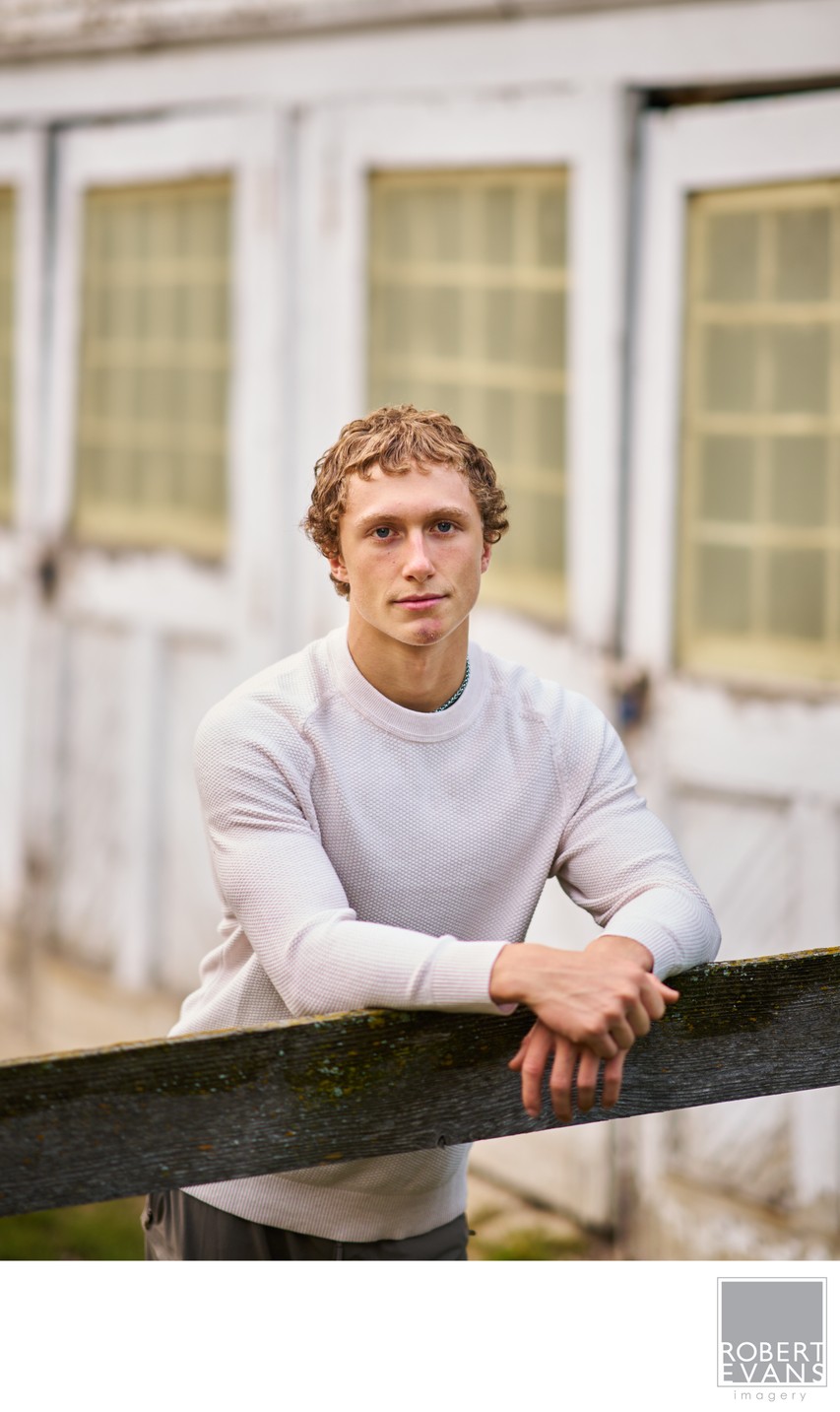 A young man in a white sweater leans against a fence beside a house.