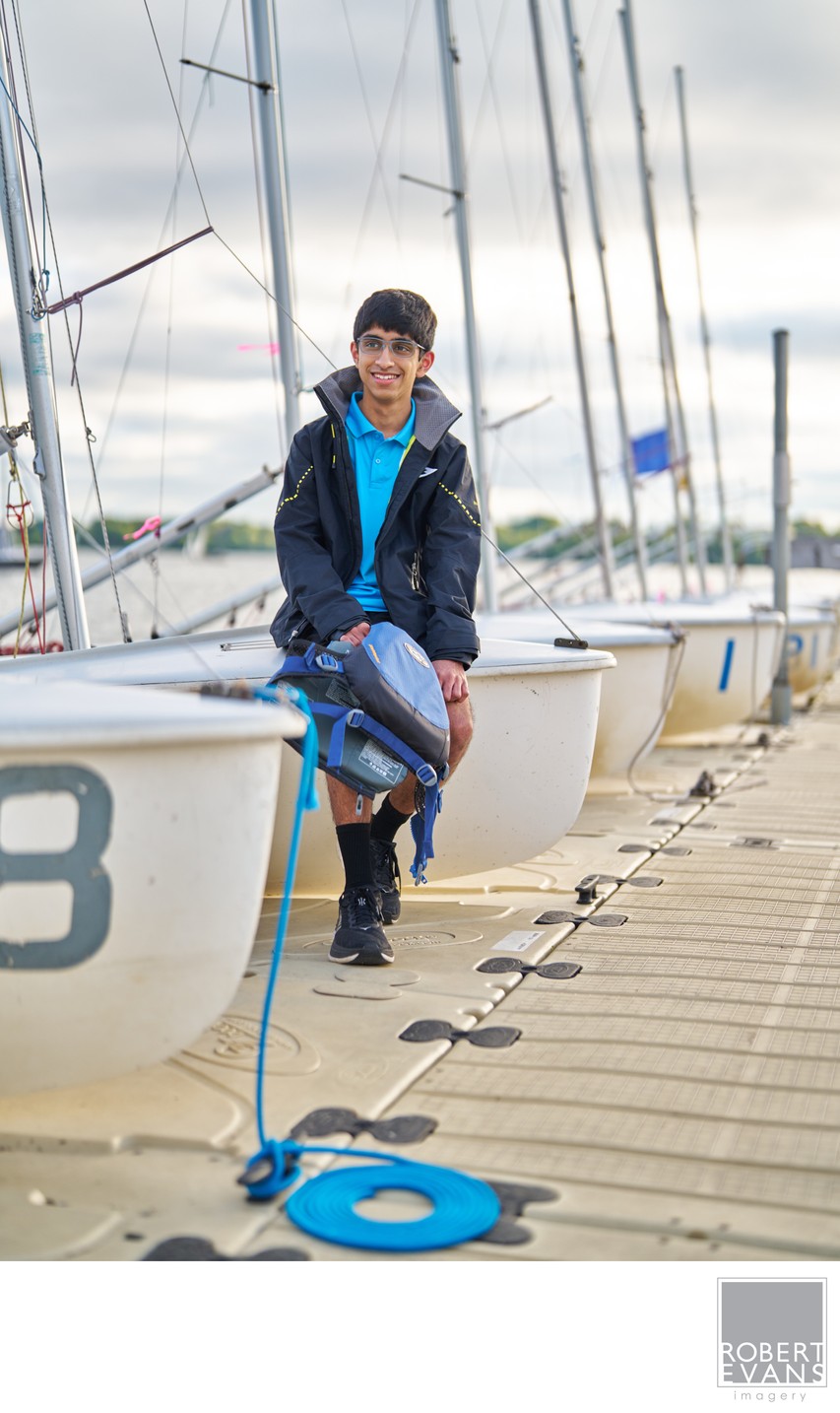 A young man sits on a dock beside sailboats and mooring ropes.