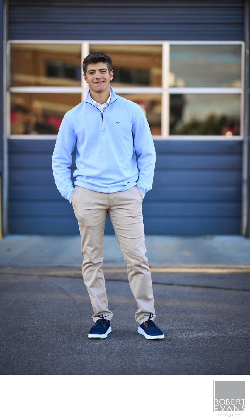A young man in a blue shirt and khaki trousers stands by a blue garage.