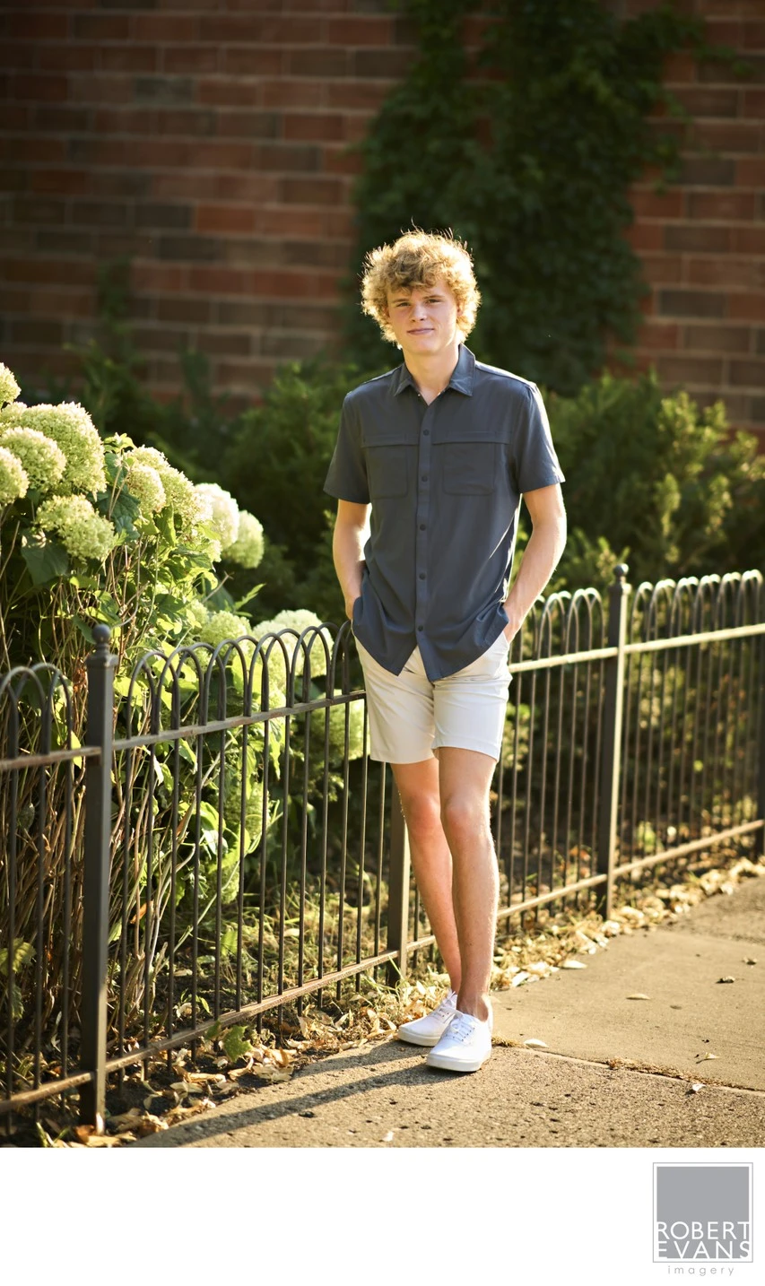 A young man in a polo shirt and shorts stands at a garden fence beside brickwork.