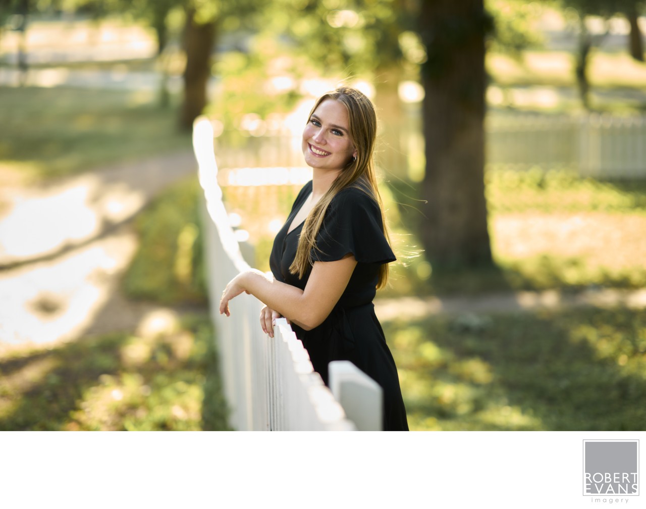 A young woman in a black dress stands beside a white fence.