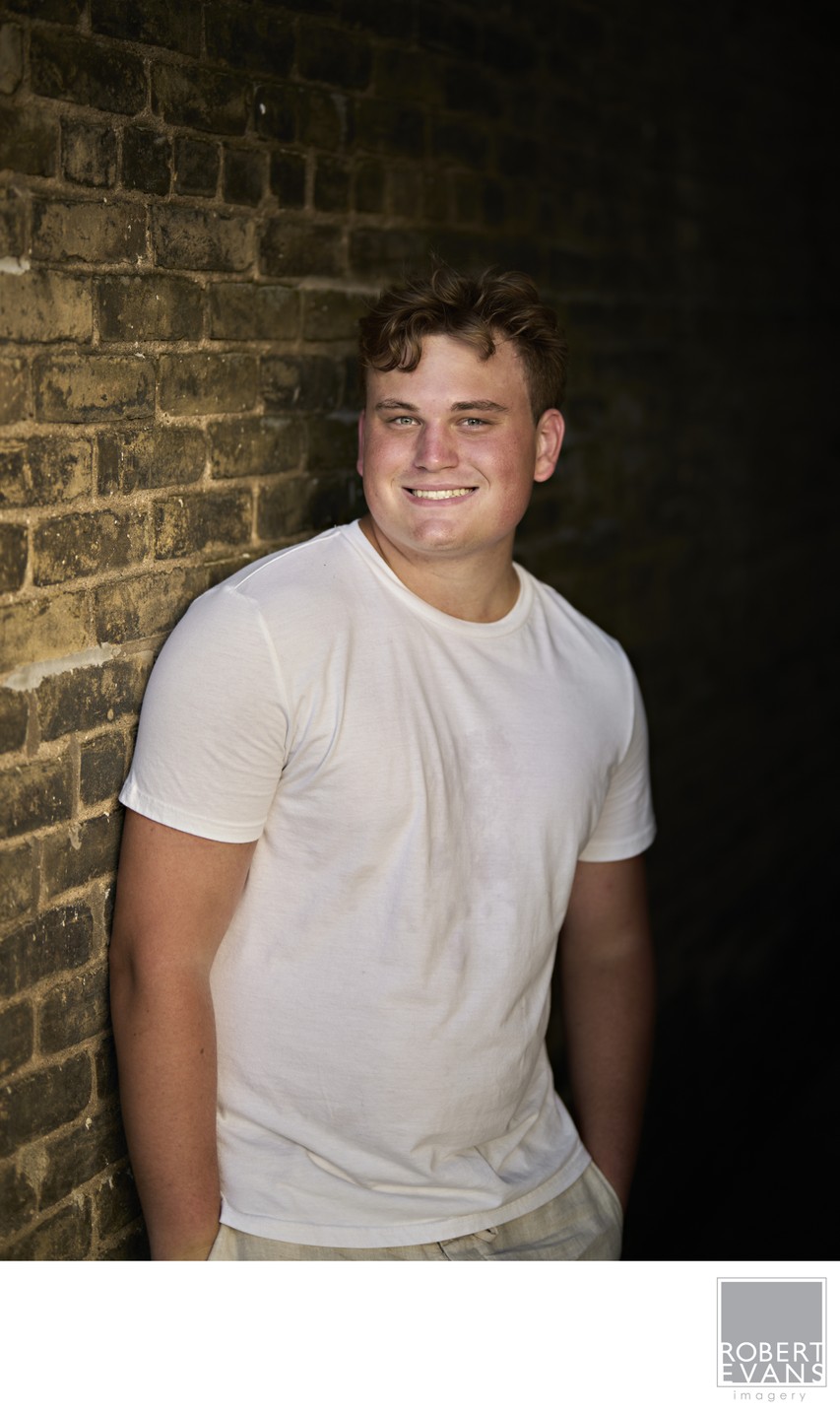 A young man in a white shirt poses against a dark brick background.