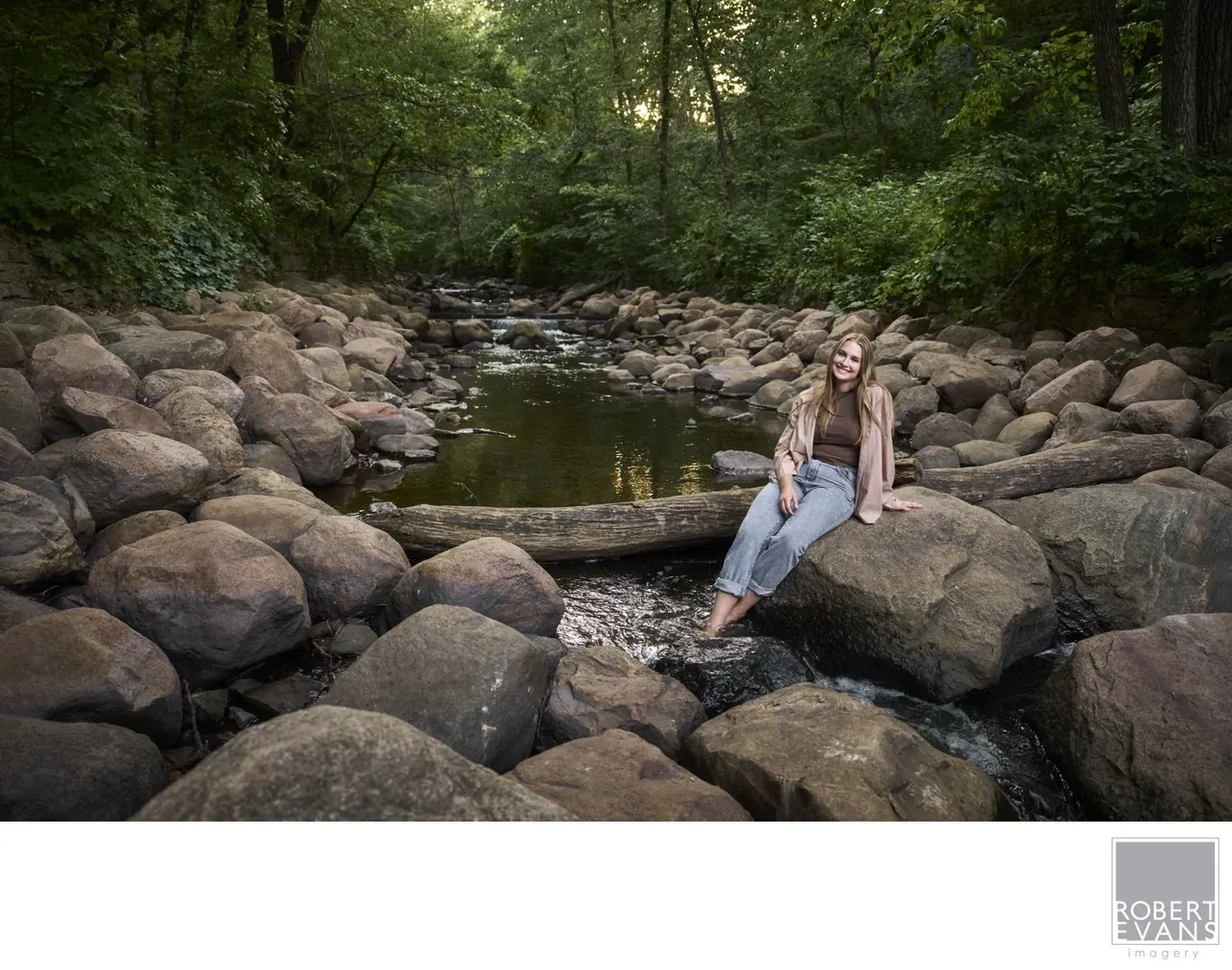 A young woman sits on rocks beside a woodland stream.