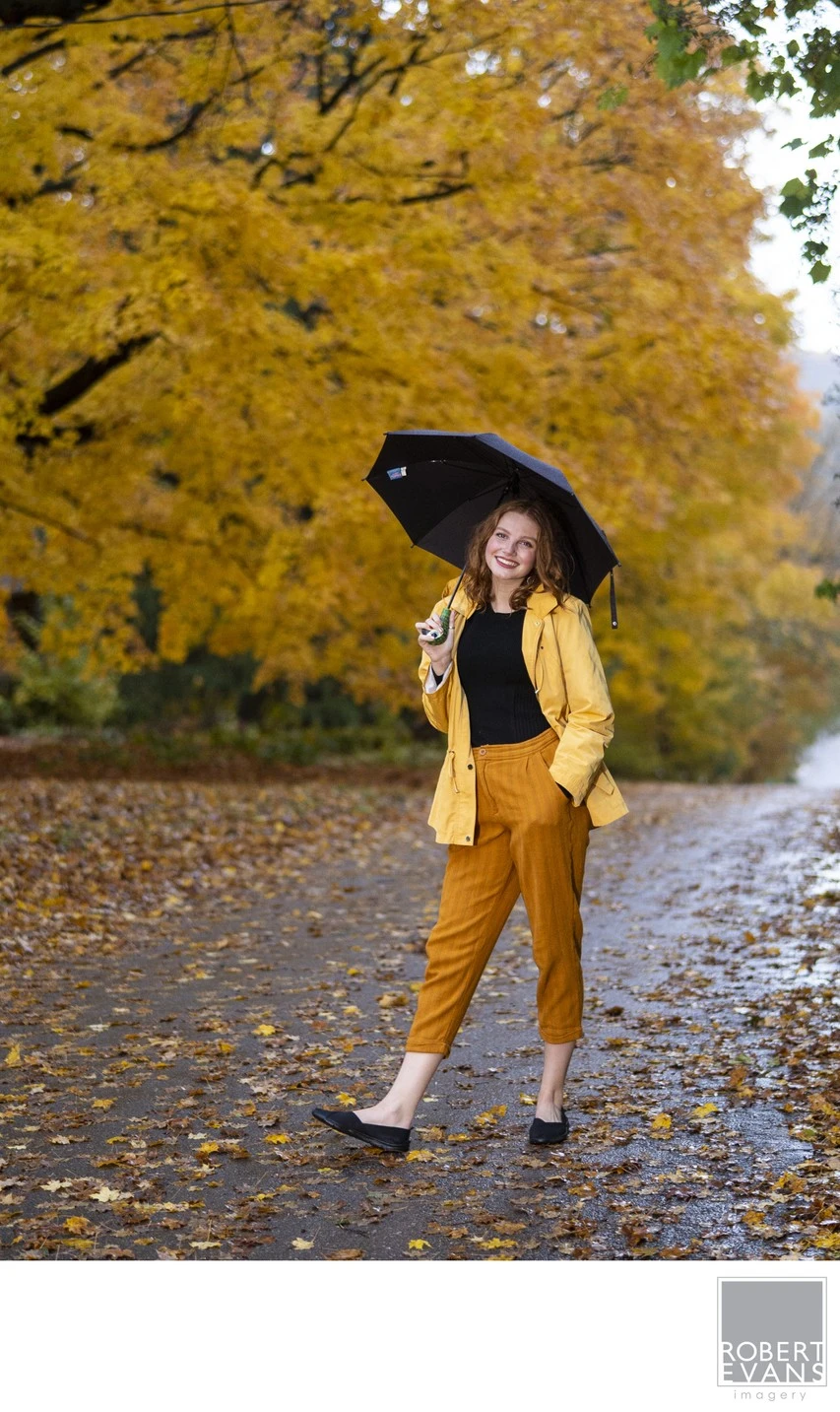 A young woman in a yellow raincoat holds a black umbrella beneath autumn trees.